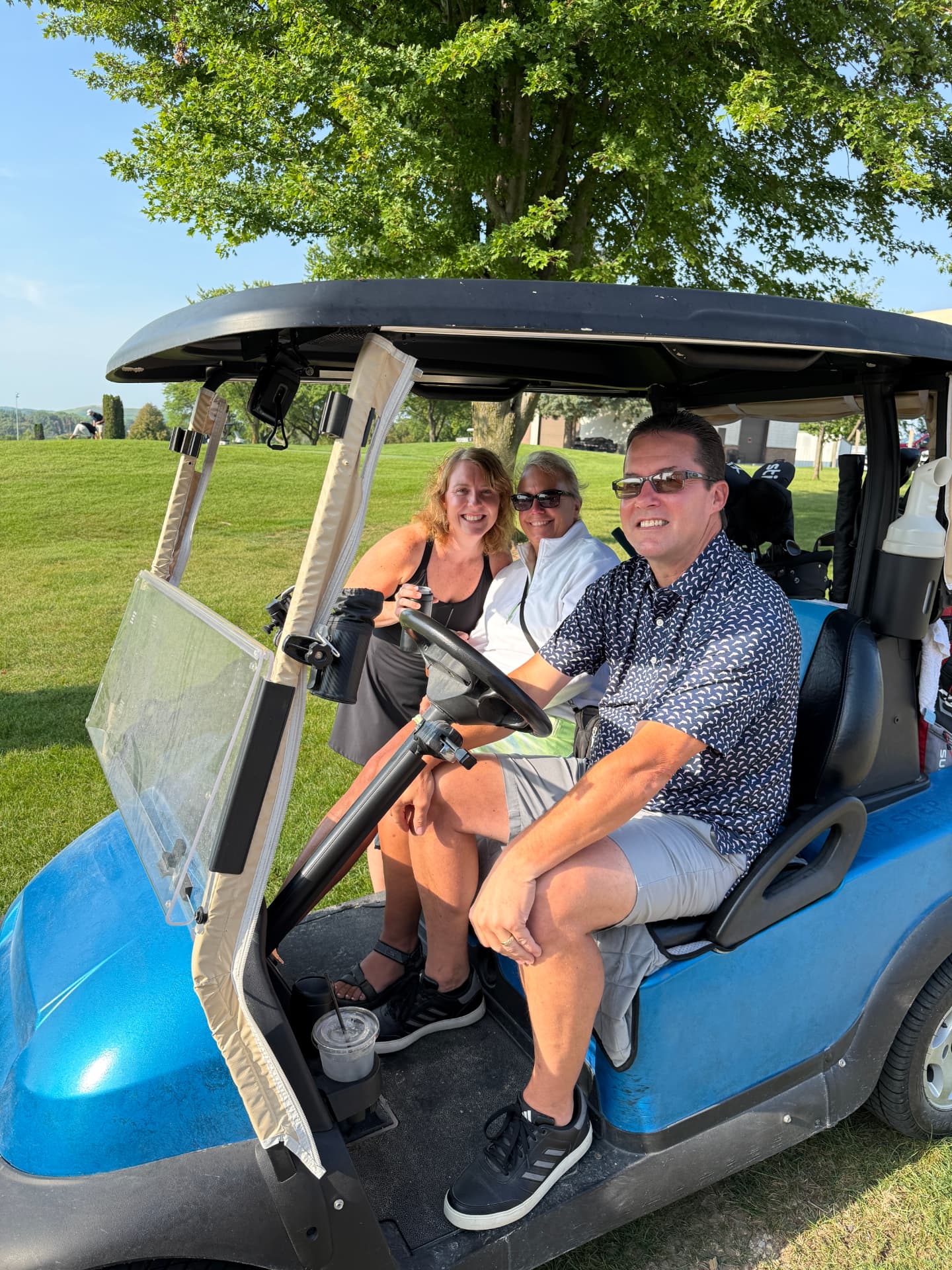 Three adults laughing beside a blue golf cart on a tree-lined fairway, one holding a beverage can.
