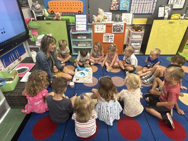A teacher kneeling beside a circle of young children as one child in a glove touches food coloring floating in a plate of milk during a classroom experiment