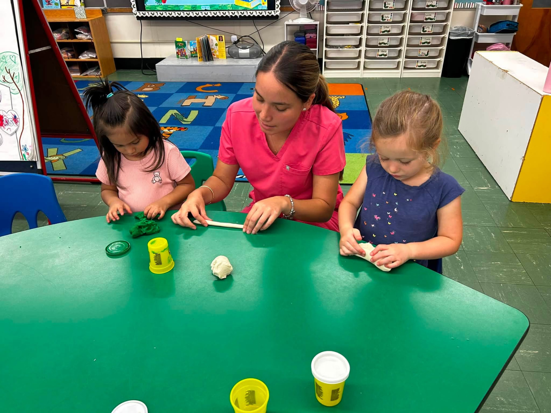 A 3K teacher rolls playdough with two children at a classroom table