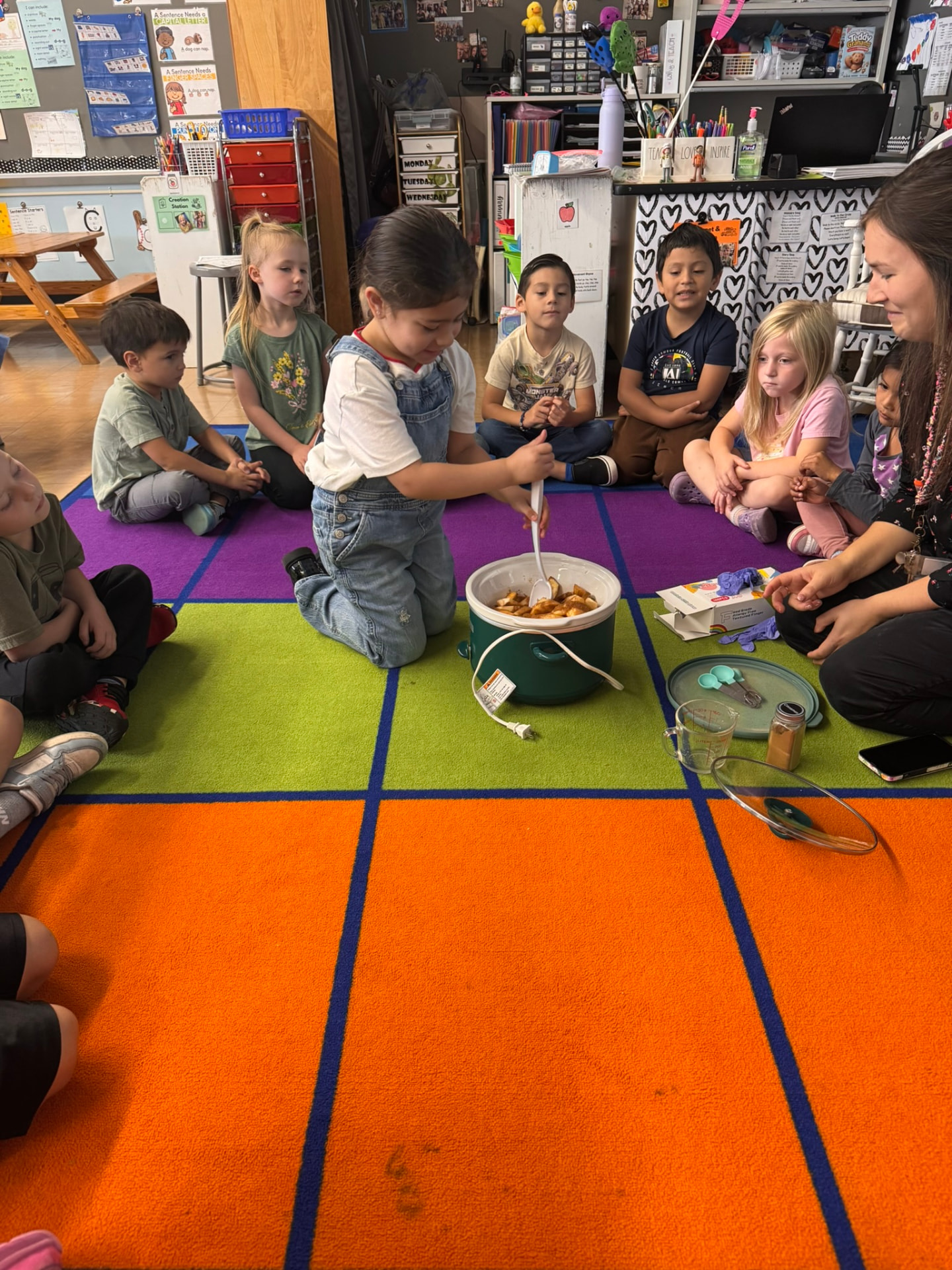 A young girl in denim overalls kneels to stir diced apples in a green slow cooker while classmates and their teacher sit around a bright classroom rug