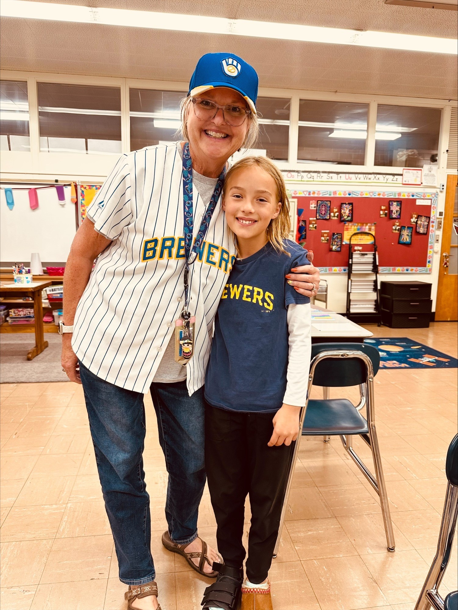 An adult in a Brewers baseball jersey and cap stands with her arm around a smiling young girl wearing a matching Brewers shirt in a classroom