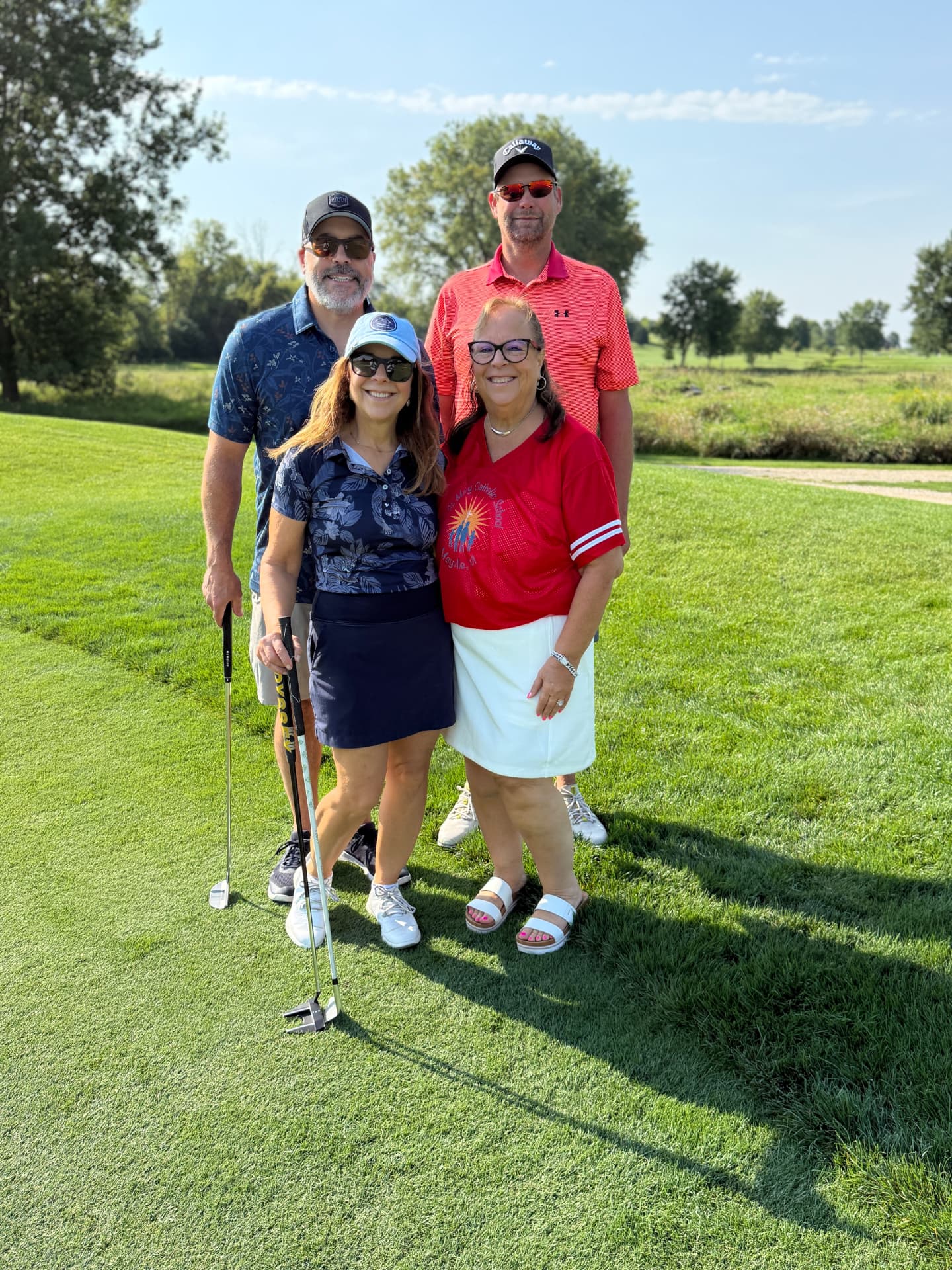 A foursome of four golfers posing together on a sunlit fairway, one wearing a red St. Mary Catholic School Mayville shirt.
