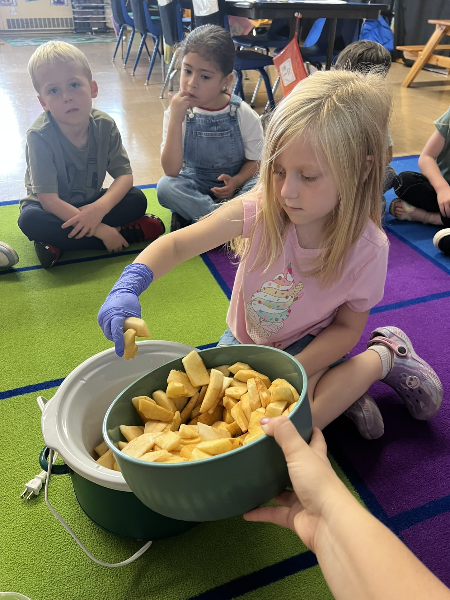 Students make applesauce together in the classroom, adding apple slices to a slow cooker