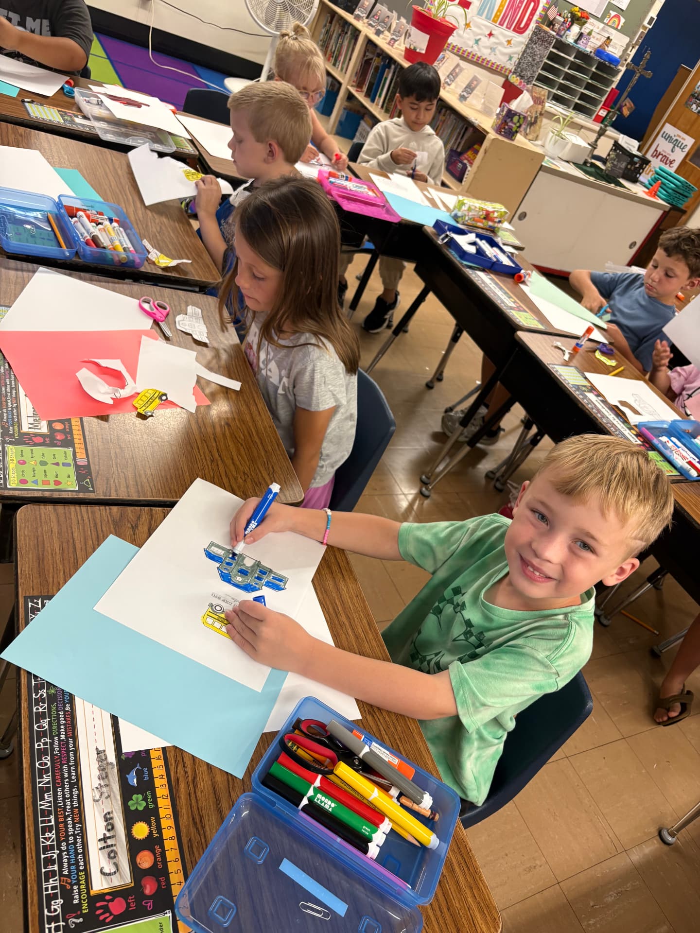 A young boy smiles while coloring a paper craft at his desk as classmates work nearby