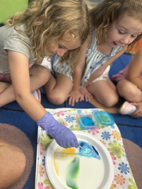 Two young students lean over a paper plate of swirling colors, dipping a cotton swab into food coloring during a classroom experiment