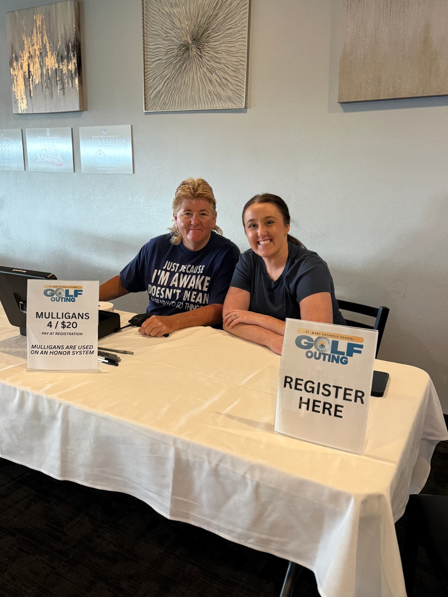 Two volunteers smiling behind the St. Mary Catholic School Golf Outing registration table, with a Register Here sign and a Mulligans sign.