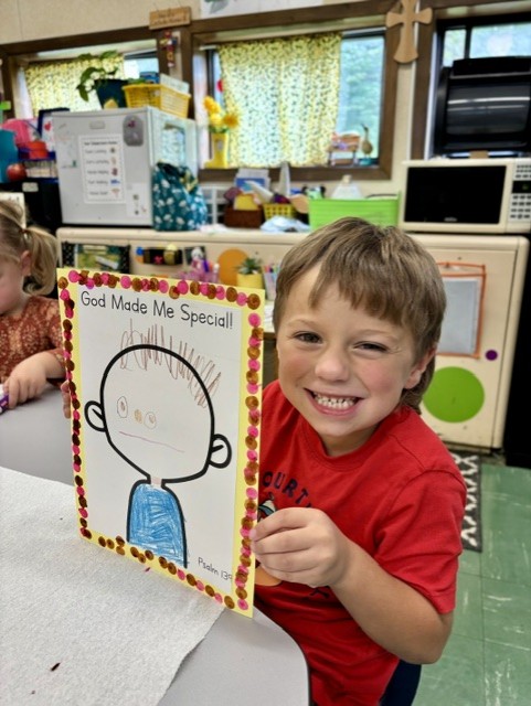 A smiling student holds up his 'God Made Me Special!' artwork decorated with dot stickers