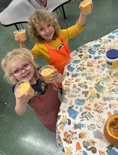 Two smiling children hold up cups of pumpkin puree at a classroom sensory table