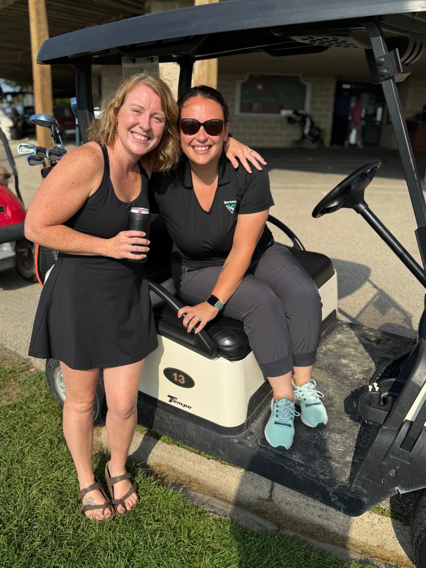 Two women smiling at the camera, one standing with her arm around the other who is seated in a cream golf cart at the cart barn.